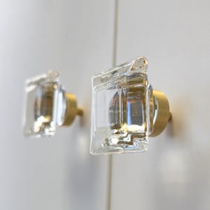 May include: Two square cabinet knobs featuring clear, thick glass fronts with a subtle rippled texture. Each knob is mounted on a circular, brushed gold-toned metal base, providing a contrast between the transparent glass and the metallic finish.