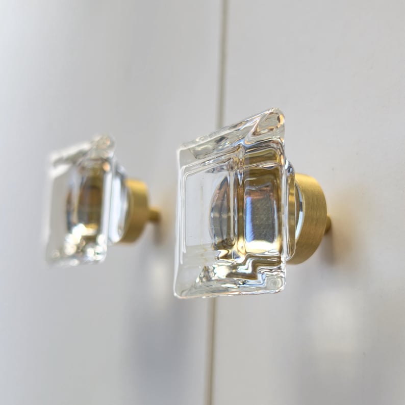 May include: Two square cabinet knobs featuring clear, thick glass fronts with a subtle rippled texture. Each knob is mounted on a circular, brushed gold-toned metal base, providing a contrast between the transparent glass and the metallic finish.