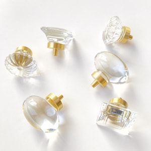 May include: A collection of six cabinet knobs featuring clear glass or acrylic tops in various shapes, including round, oval, and square designs. Each knob is attached to a circular brushed gold metal base with a central mounting post.