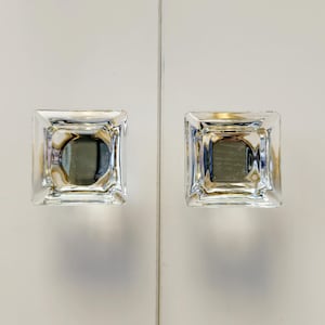 May include: Two square cabinet knobs made of clear, faceted glass. Each knob features a geometric, multi-layered design with beveled edges that create a prismatic effect, reflecting light through the transparent material to show depth.