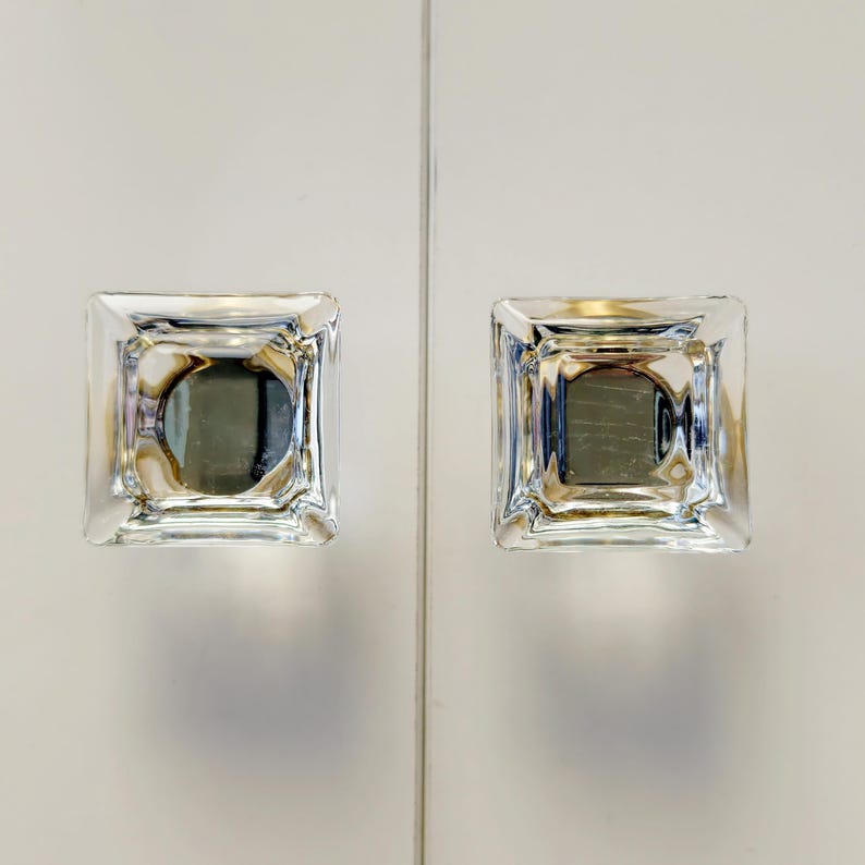 May include: Two square cabinet knobs made of clear, faceted glass. Each knob features a geometric, multi-layered design with beveled edges that create a prismatic effect, reflecting light through the transparent material to show depth.