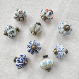 May include: A collection of eight ceramic drawer knobs with a fluted, pumpkin-like shape. Each knob features hand-painted floral or leaf patterns in various colours including blue, green, and orange, finished with a central antique brass metal base.