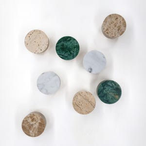 Puede incluir: Una colección de ocho pomos de piedra circulares dispuestos en un patrón disperso. El conjunto incluye piezas con diversas texturas y colores, que presentan travertino beige, mármol verde oscuro, mármol blanco con vetas grises y piedra pulida marrón.
