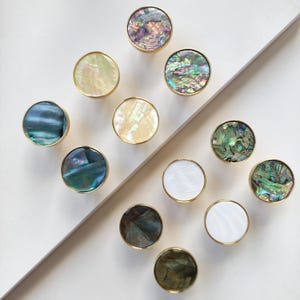 May include: A collection of twelve circular cabinet knobs featuring iridescent abalone shell and smooth white mother-of-pearl inlays. Each knob is encased in a thin gold-toned metal rim, displaying a variety of natural colours including deep blues, greens, purples, and creamy white tones.
