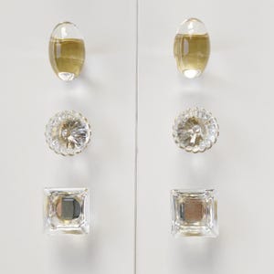 May include: A set of six cabinet knobs arranged in two vertical columns. The top pair are smooth oval shapes in a translucent amber tone. The middle pair are round with a faceted, starburst-like texture. The bottom pair are clear square knobs.