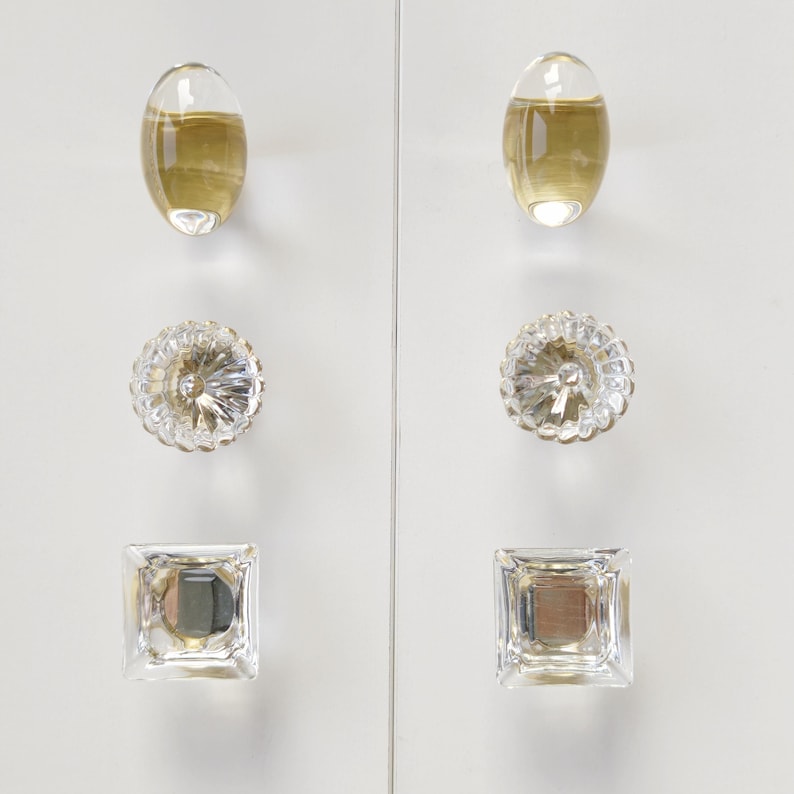 May include: A set of six cabinet knobs arranged in two vertical columns. The top pair are smooth oval shapes in a translucent amber tone. The middle pair are round with a faceted, starburst-like texture. The bottom pair are clear square knobs.