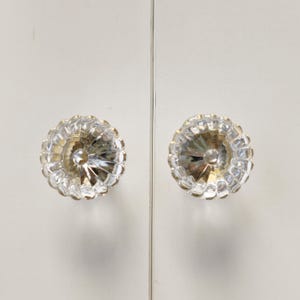 May include: Two circular, clear glass cabinet knobs featuring a faceted, sunburst-like pattern with radiating ridges. Each knob has a smooth, rounded center and a scalloped outer edge, mounted on a flat, white, vertical surface with a central seam.