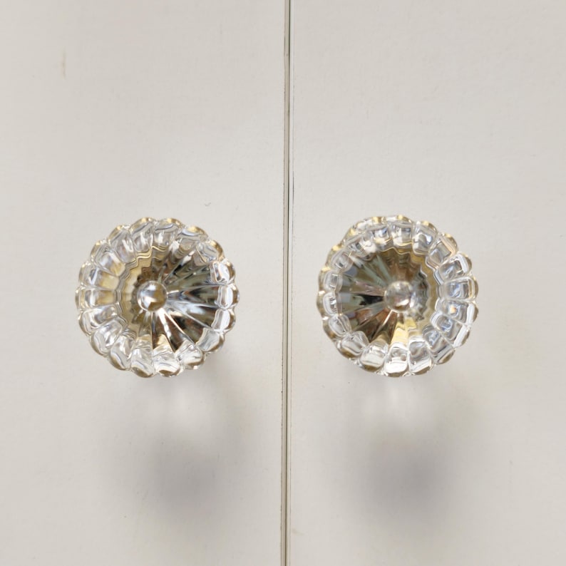 May include: Two circular, clear glass cabinet knobs featuring a faceted, sunburst-like pattern with radiating ridges. Each knob has a smooth, rounded center and a scalloped outer edge, mounted on a flat, white, vertical surface with a central seam.