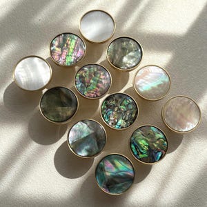 May include: A collection of eleven circular cabinet knobs featuring iridescent abalone shell and smooth white mother-of-pearl inlays set within gold-toned metal rims, arranged in a diamond pattern on a light-colored surface.
