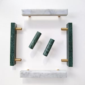 May include: A collection of cabinet hardware including two white marble rectangular pulls, two dark green marble rectangular pulls, and two cylindrical green marble knobs, all featuring brushed brass metal mounting posts.