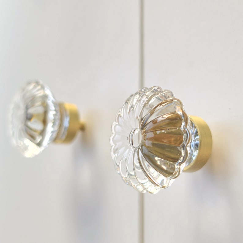 May include: Two circular cabinet knobs featuring clear, fluted glass fronts shaped like flower petals. Each knob is mounted on a cylindrical, brushed gold-toned metal base, providing a contrast between the transparent glass and the metallic finish.