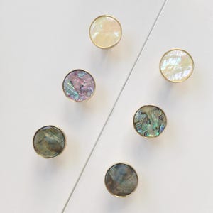 Puede incluir: Seis pomos circulares para armarios con bordes de metal dorado y centros decorativos incrustados. Las incrustaciones presentan variados patrones iridiscentes, nacarados y veteados en tonos blanco, crema, morado, azul, verde y gris oscuro.