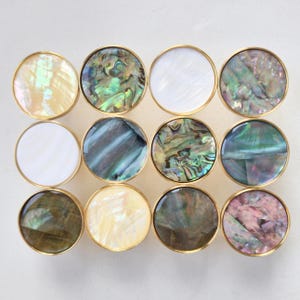 May include: A collection of twelve circular cabinet knobs arranged in a grid. Each knob features a polished brass rim surrounding a unique inlay of iridescent shell, including white, cream, green, blue, and purple tones with natural patterns.