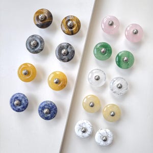 May include: A collection of fourteen circular cabinet knobs made from various polished stones and minerals, each featuring a central silver-toned metal screw head. The knobs display diverse colours including tiger eye, grey, yellow, blue, pink, green, and white.