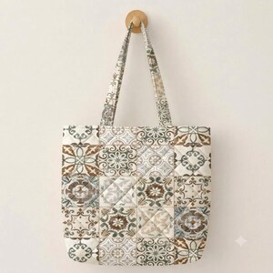 May include: A quilted tote bag featuring a repeating grid pattern of square tiles. The design displays various intricate floral and geometric motifs in muted shades of brown, beige, gray, and white. The bag includes two matching fabric shoulder straps.