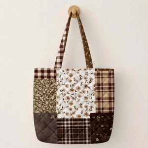 May include: A quilted tote bag featuring a patchwork design of brown and white fabrics. The pattern includes floral prints, gingham checks, and plaid squares. The bag has two long shoulder straps and is suspended from a wooden wall hook.