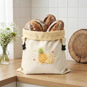 May include: A cream-colored canvas bread bag with a pineapple and flower graphic print. The bag features a light yellow interior lining, black plastic side buckles, and is filled with three round, crusty loaves of artisan sourdough bread.