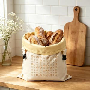 May include: A cream-colored fabric bread bag with a light yellow interior lining and black plastic side buckles. The exterior features a repeating pattern of small brown floral and geometric icons. Several crusty loaves of bread fill the bag.