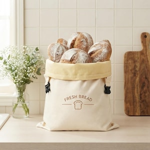 May include: A cream-colored canvas bread bag with a folded top edge, featuring the text "FRESH BREAD" and a small bread icon printed in brown. The bag is filled with several round, crusty loaves of bread and has black plastic side buckles.