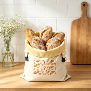 May include: A cream-colored fabric bread bag with a yellow interior lining and black side buckles. The exterior features a brown graphic print of various bread types including loaves, croissants, and wheat stalks, filled with several crusty bread loaves.