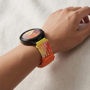 Könnte beinhalten: Eine Smartwatch mit schwarzem, rundem Gehäuse und orangefarbenem Zifferblatt mit Farbverlauf. Das gewebte Stoffarmband zeigt ein Zickzackmuster in Gelb, Helllila und Orange und wird mit einer leuchtend orangefarbenen Kunststoffschnalle befestigt.
