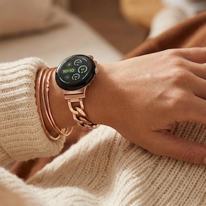 May include: A rose gold smartwatch with a circular display showing the time 10:10 and various health metrics. The watch features a metallic chain-link band and is worn alongside three thin, matching rose gold bangles on a wrist.