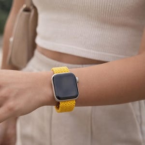 May include: A silver smartwatch featuring a rectangular black screen and a bright yellow braided elastic watch band. The watch case has a metallic finish, and the strap displays a consistent woven texture that wraps around the wrist.