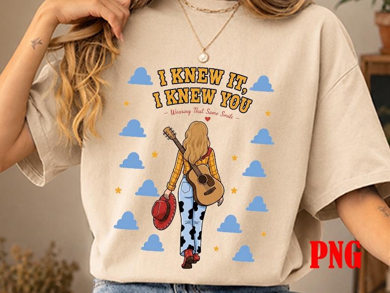 May include: A beige t-shirt featuring a graphic of a person with blonde hair, wearing a yellow plaid shirt and cow-print pants, carrying a guitar and a red hat. The text reads I KNEW IT, I KNEW YOU and Wearing That Same Smile, surrounded by blue clouds.