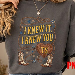 May include: A charcoal gray short-sleeve t-shirt featuring a graphic print with the text I KNEW IT, I KNEW YOU, TS inside a heart, surrounded by a lasso, blue stars, and two brown and white cowboy boots with intricate western-style detailing.