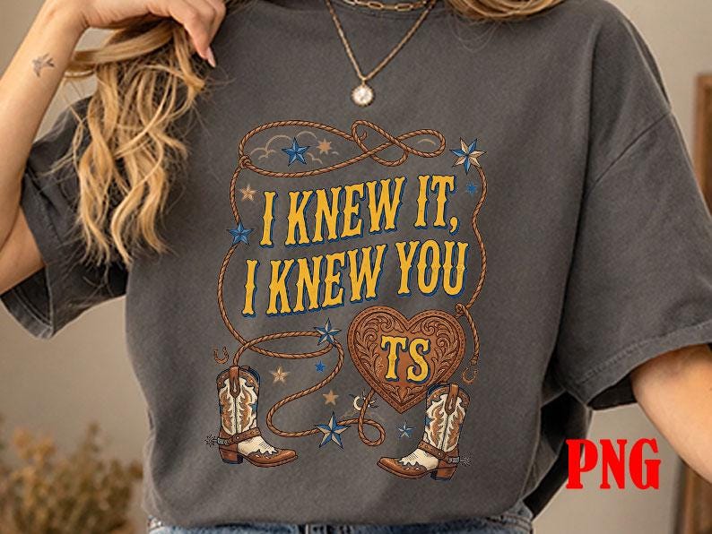 May include: A charcoal gray short-sleeve t-shirt featuring a graphic print with the text I KNEW IT, I KNEW YOU, TS inside a heart, surrounded by a lasso, blue stars, and two brown and white cowboy boots with intricate western-style detailing.