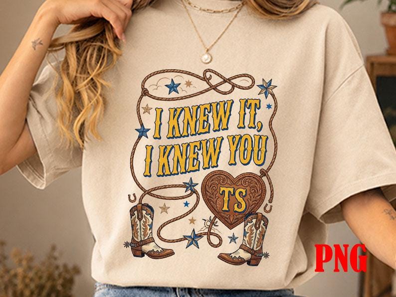 May include: A beige short-sleeve t-shirt featuring a graphic print with a lasso, two cowboy boots, and a heart shape containing the letters TS. The text reads I KNEW IT, I KNEW YOU. The design includes blue stars and brown rope accents.