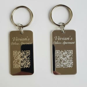 May include: Two rectangular silver-toned metal keychains with a polished finish. Each keychain features the engraved text 'Vivian's Deluxe Apartment' above a square QR code. A circular metal split ring is attached to the top of each tag.