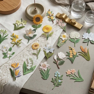 May include: A collection of various embroidered flower patches featuring diverse botanical designs including sunflowers, lilies, daffodils, and bird of paradise flowers, each with detailed green leaves and stems, arranged on a white lace-trimmed fabric.