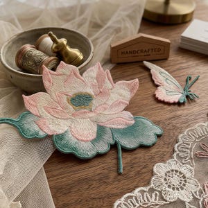 Könnte beinhalten: Ein gestickter Lotusblüten-Aufnäher in Rosa- und Petroltönen, begleitet von einem kleineren Schmetterlings-Aufnäher in passenden Farben. Ein Stück weiße Spitze mit Blumenstickerei ist sichtbar, neben einer Schale mit Garnrollen und einem Messingwerkzeug.