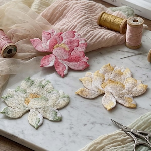 Könnte beinhalten: Drei bestickte Blumenaufnäher in Pink, Gelb und Weiß mit grünen Akzenten. Die Aufnäher weisen detaillierte Stickereien auf und sind zusammen mit hölzernen Garnrollen, Nähnadeln, Stoffresten und einer Stickschere aus Metall angeordnet.