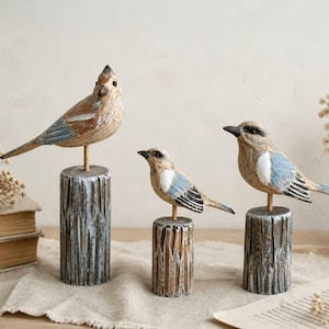 Peut inclure: Trois figurines d'oiseaux en bois sculpté perchées sur des socles cylindriques en forme de bûche. Chaque oiseau présente des détails peints en brun, blanc et bleu sur les ailes et le corps. Les oiseaux sont fixés sur de fines tiges en bois.