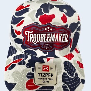 Puede incluir: Una gorra de camionero de cinco paneles con un patrón de camuflaje en blanco, azul marino y rojo. El frente muestra un parche rojo y blanco con el texto TROUBLEMAKER. Una pegatina cuadrada en la visera dice EST. 1970 R 112PFP PRINTED 5 PANEL OSFM.