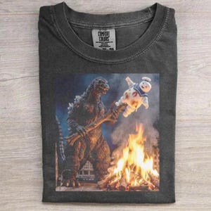 May include: A charcoal gray crew neck t-shirt featuring a graphic print of Godzilla holding a stick with a toasted Stay Puft Marshmallow Man over a campfire. The inner neck label displays the text Comfort Colors and size information.