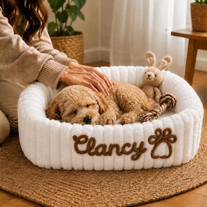 May include: A white, ribbed, rectangular pet bed featuring the embroidered brown name Clancy and a brown paw print. Inside the bed, a light brown curly-haired puppy is sleeping next to a small, crocheted, tan-coloured rabbit toy.