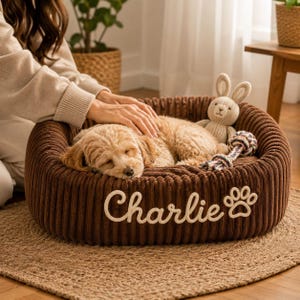 Puede incluir: Una cama redonda para mascotas de pana marrón con el nombre Charlie bordado y un icono de huella en la parte frontal. Dentro de la cama, un cachorro de color claro duerme junto a un pequeño conejo de punto y un juguete de cuerda.