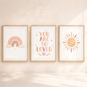 Könnte beinhalten: Drei gerahmte Wandbilder in hellen Holzrahmen. Das linke Bild zeigt einen Aquarell-Regenbogen mit Wolken. Das mittlere Bild enthält den Text "YOU ARE SO LOVED" mit Sternen und einem Herz. Das rechte Bild zeigt eine Illustration einer lächelnden Sonne.