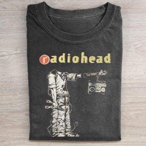 May include: A charcoal gray crew neck t-shirt featuring the text Radiohead in yellow letters next to a red circle. Below the text is a black and white graphic illustration of a headless figure holding a boombox with a cord and plug attached.