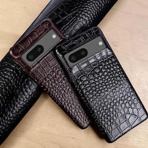 May include: Two smartphone cases featuring a textured crocodile skin pattern. One case is dark brown and the other is black. Both cases have a rectangular camera cutout at the top with two circular lenses and a small flash visible on the back surface.