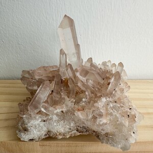 May include: A cluster of clear quartz crystals featuring a prominent, tall central point surrounded by numerous smaller, jagged crystalline formations. The mineral specimen displays translucent, pale pinkish-white hues and sharp, geometric facets.