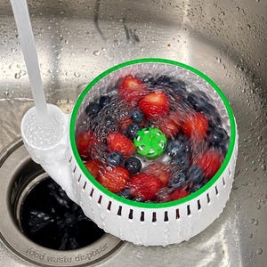 May include: A white circular fruit washing colander with a green rim and a small green center spinner, filled with fresh red strawberries and dark blue blueberries being rinsed under a stream of water in a stainless steel kitchen sink.