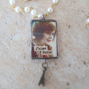 K&ouml;nnte beinhalten: Eine silberne Halskette mit einem rechteckigen Anh&auml;nger, der ein Vintage-Bild einer Frau mit dem Text "J'aime Paris" zeigt. Der Anh&auml;nger ist an einer silbernen Kette mit einem kleinen Eiffelturm-Anh&auml;nger befestigt.