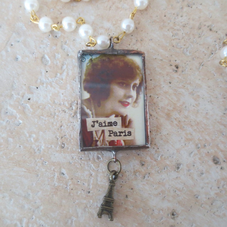 K&ouml;nnte beinhalten: Eine silberne Halskette mit einem rechteckigen Anh&auml;nger, der ein Vintage-Bild einer Frau mit dem Text "J'aime Paris" zeigt. Der Anh&auml;nger ist an einer silbernen Kette mit einem kleinen Eiffelturm-Anh&auml;nger befestigt.