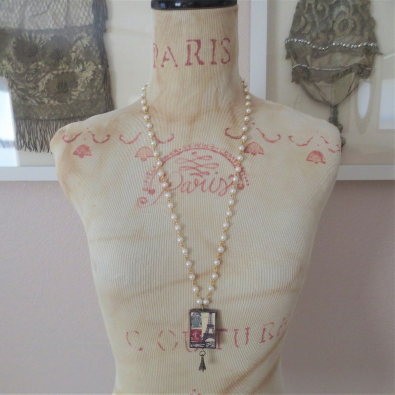 K&ouml;nnte beinhalten: Eine wei&szlig;e Perlenkette mit einer goldenen Kette und einem rechteckigen Anh&auml;nger mit einer Collage von Pariser Sehensw&uuml;rdigkeiten, darunter der Eiffelturm. Der Anh&auml;nger hat einen kleinen Anh&auml;nger, der daran h&auml;ngt. Die Kette wird auf einer wei&szlig;en Schaufensterpuppe mit dem Wort "PARIS" auf der Brust pr&auml;sentiert.