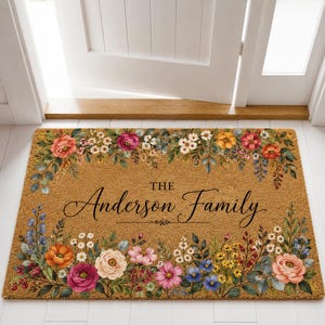Puede incluir: Un felpudo rectangular de fibra de coco color canela con un borde floral vibrante de flores rosas, naranjas, amarillas y azules con follaje verde. El centro muestra el texto negro "THE Anderson Family" en fuentes serif y cursiva.