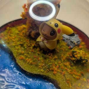 Op de afbeelding: Een doorzichtige bolvormige vitrine met een miniatuur diorama van een klein bruin wezen met een gele Pikachu-capuchon. De scène bevat een blauw watergedeelte, groen en oranje bodembedekking, een boom met textuur en een grijze rots.